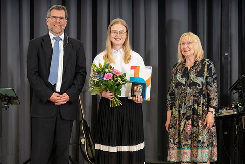Prof. Dr. Litschen (links), die Preisträgerin des Fakultätspreises 2025, Philomena Mercedes Susan Finke-Fricke, und die stellvertretende Bürgermeisterin Elke Wesche-Möller lächeln in die Kamera. Die Preisträgerin in weißer Bluse und dunklem Rock hält ihre Auszeichnung und einen bunten Blumenstrauß. (öffnet Vergrößerung des Bildes)