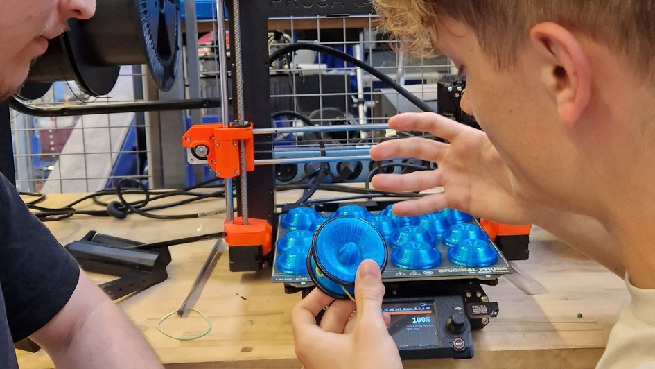 Zwei Studierende halten ein fertiges Yoyo. Im Hintergrund ist ein 3-D-Drucker zu sehen, auf dem weitere Yoyo-Schalen im Druck sind. 