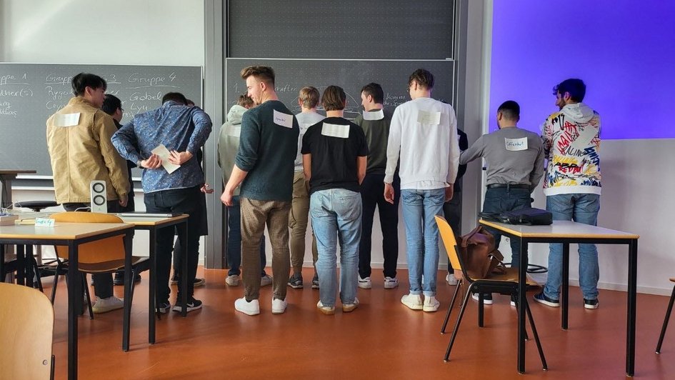 Zu sehen ist eine Gruppe Studierender vor einer Tafel, die diskutieren und sich unterhalten