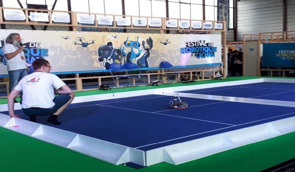 Ein kleiner Roboter bewegt sich über eine abgegrenzte blaue Fläche. Im Hintergrund sind zwei Personen zu sehen, neben ihnen der Schriftzug "Festival de Robotique de Cachan"