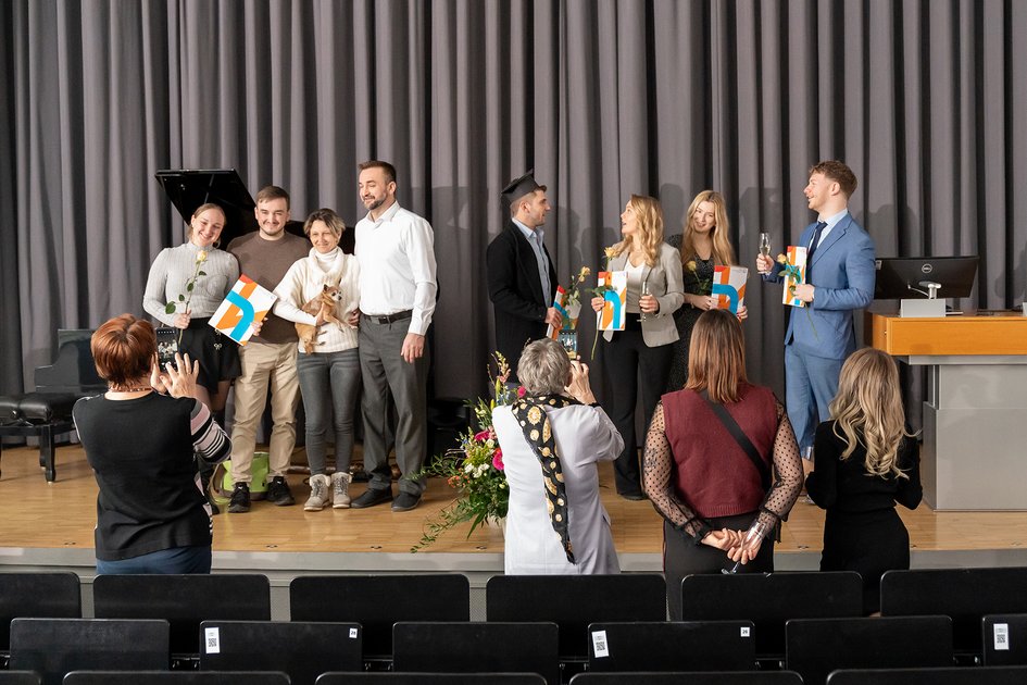 Blick auf die Bühne, vor der Angehörige stehen, welche Fotos von den Absolvent:innen machen, die ihre Urkunden halten.