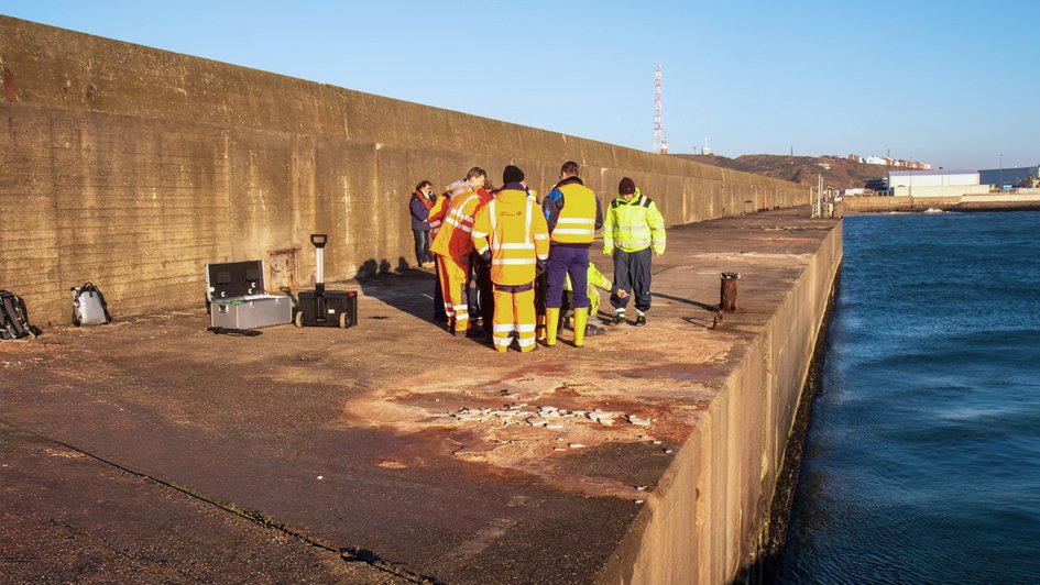 Sechs Studierende in Baukleidung stehen auf einer Hafenmole in Helgoland und machen Messungen. Links ist eine Betonmauer, rechts das Hafenbecken.
