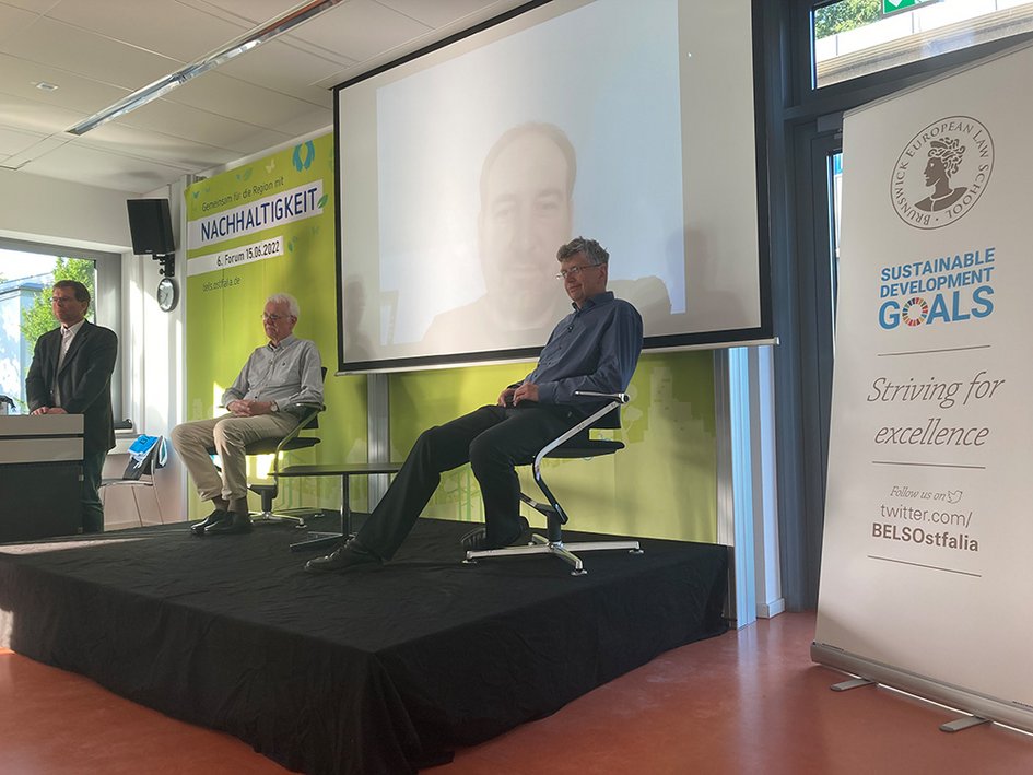 Prof. Litschen steht links am Rednerpult, Hans-Heinrich Schmidt-Kanefendt, Benjamin Volz (digital zugeschaltet), und Prof. Dr. Jürgen Kuck diskutieren miteinander, Benjamin Volz ist auf einer Leinwand zu hinter der Bühne zu sehen.