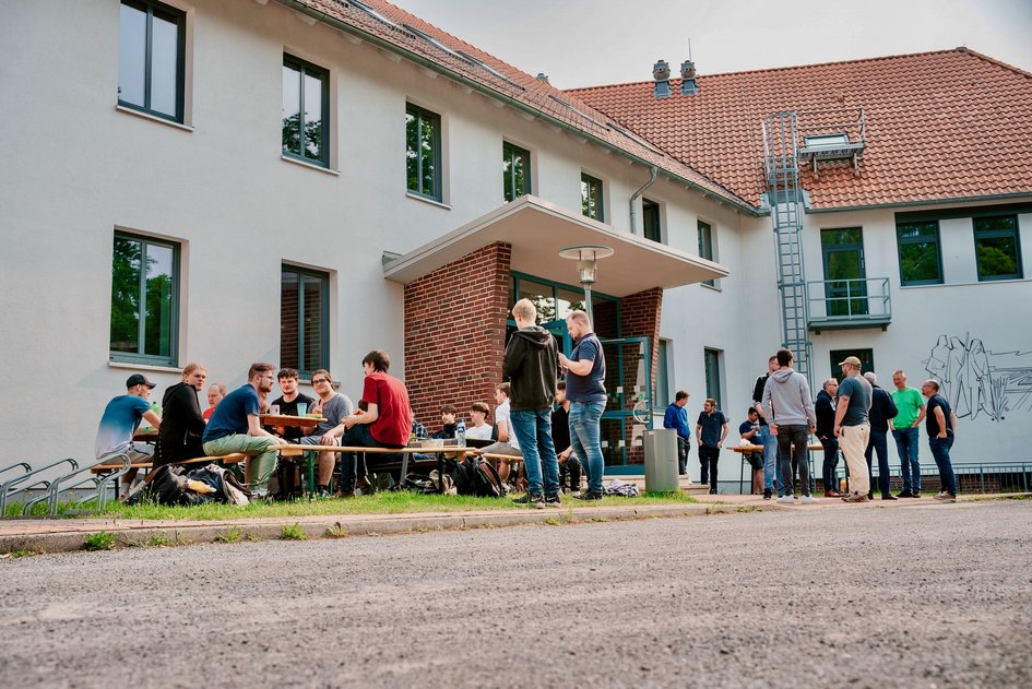 Studierende und Lehrende des Studienganges Angewandte Informatik stehen und sitzen an Tischen und vor dem Gebäude A während eines gemeinsamen Frühstücks.