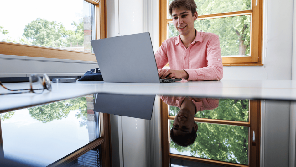 Dekorativ: Person sitzt an einem Tisch vor einem Laptop und nutzt einen mobilen Router für Online-Studium in einem hellen Raum mit großen Fenstern. (öffnet Vergrößerung des Bildes)
