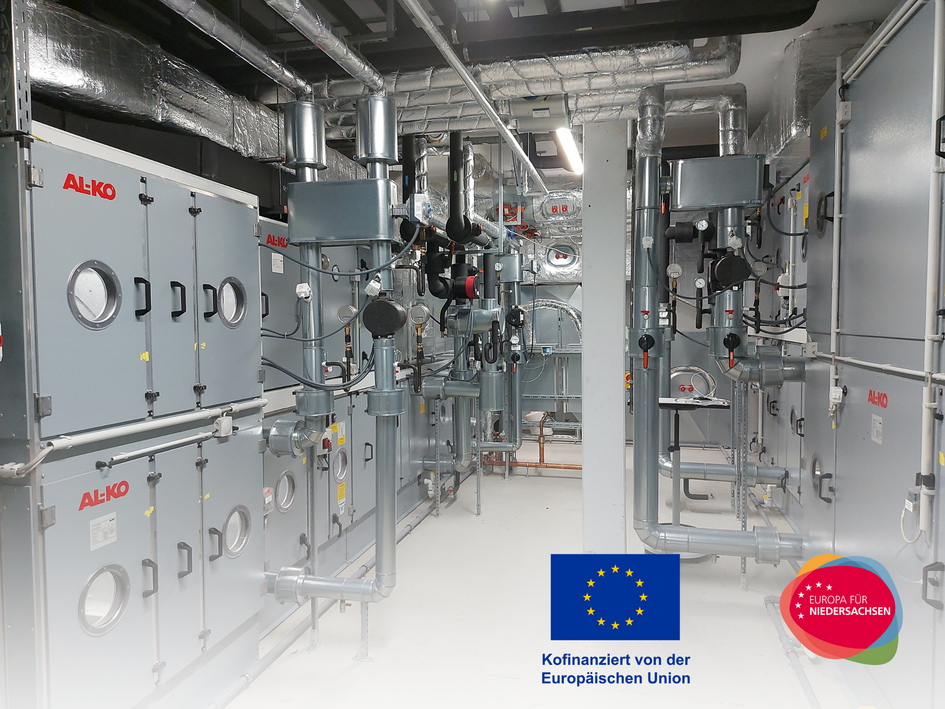 Das Bild zeigt eine moderne raumlufttechnische Anlage (RLT-Anlage) in einem Technikraum. Mehrere silberne Lüftungskanäle verlaufen über kompakten technischen Geräten mit Anzeigeelementen. Im Vordergrund sind sauber verlegte Rohre und Kabel zu sehen. Die Aufnahme ist hell und vermittelt den Eindruck moderner Gebäudetechnik. (öffnet Vergrößerung des Bildes)