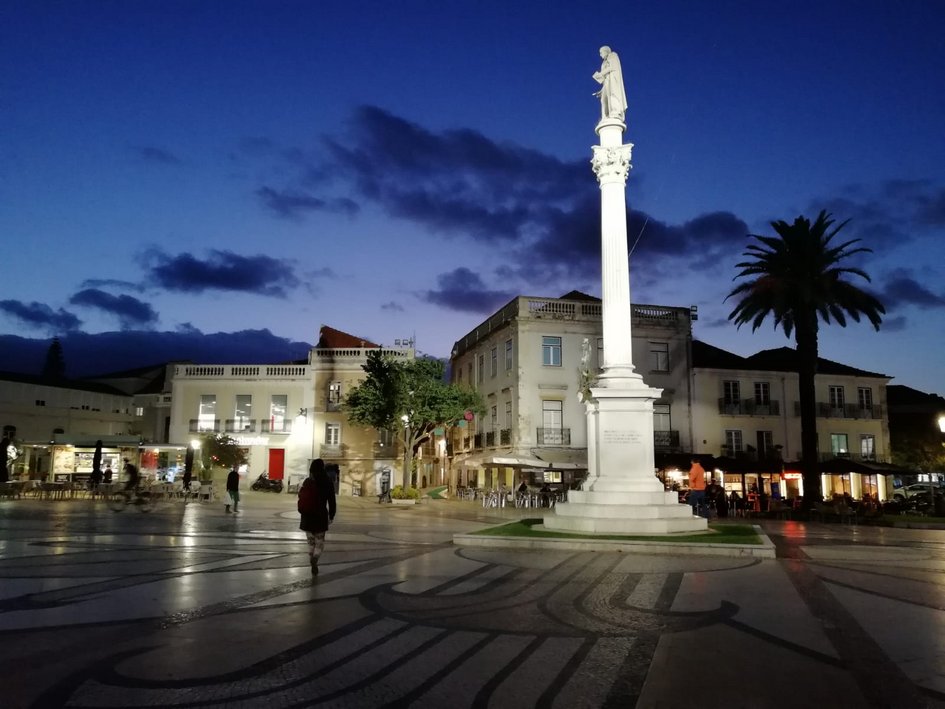A photograph of Bocage Square in the Portuguese city of Setúbal at night. The focus is placed on an illuminated statue on a white column