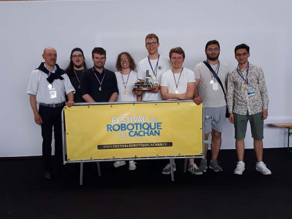 Studenten stehen hinter einem Banner mit dem Schriftzug "Festival de Robotique de Cachan". Einer der Studenten hält einen kleinen Roboter in den Händen
