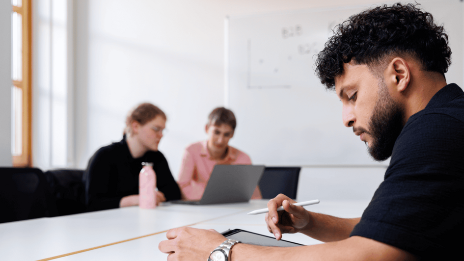 Dekorativ:Drei Studierende sitzen an einem Tisch, einer nutzt ein Tablet mit Stift, zwei weitere arbeiten am Laptop in einem hellen Raum. (öffnet Vergrößerung des Bildes)