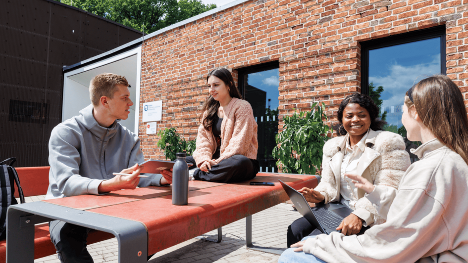 Dekorativ:Vier Studierende sitzen an einem roten Außentisch vor einem Backsteingebäude und unterhalten sich, zwei nutzen Laptop und Tablet. (öffnet Vergrößerung des Bildes)