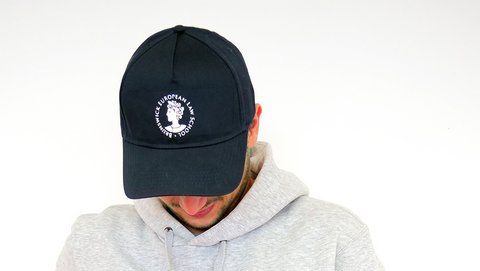 Die Cap trägt das Logo der BELS, die Aequitas, über über dem Schirm. Darum findet sich der kreisförmig angeordnete Schriftzug "Brunswick European Law School" Die Cap ist blau und das Logo und der Schriftzug sind weiß. (öffnet Vergrößerung des Bildes)