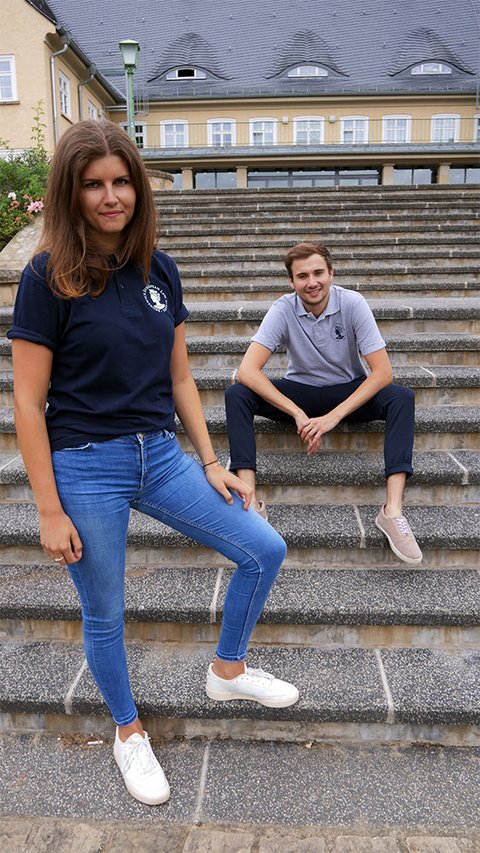 Ein junger Mann trägt das Polo-Shirt der BELS. Es ist grau melange und trägt das Logo der BELS, die Aequitas, links auf Brusthöhe. Darum findet sich der kreisförmig angeordnete Schriftzug "Brunswick European Law School". Das Logo und der Schriftzug sind blau. (öffnet Vergrößerung des Bildes)