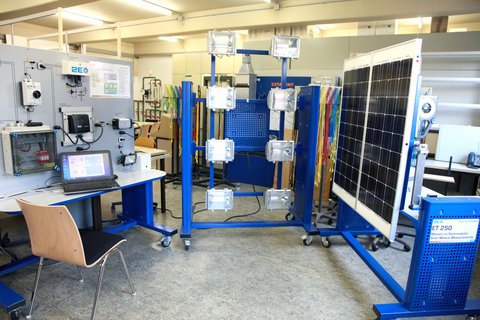 Das Bild zeigt ein im Labor für Elektrotechnik und regenerative Energietechnik aufgebautes Solarstromsystem, bestehend aus einer künstlichen Lichtquelle, einem Photovoltaikmodul und einem Arbeitsplatz mit vernetzten Komponenten des Solarstromsystems wie Laderegler, Netzwechselrichter, Akkumulator als Speicher für Strom, Zweirichtungsstromzähler sowie ein Energiemanagementsystem. Verschiedene steuerbare Verbraucher können in das System integriert werden. In einer zentralen Kommunikations- und Steuereinheit werden Daten der Komponenten erfasst.