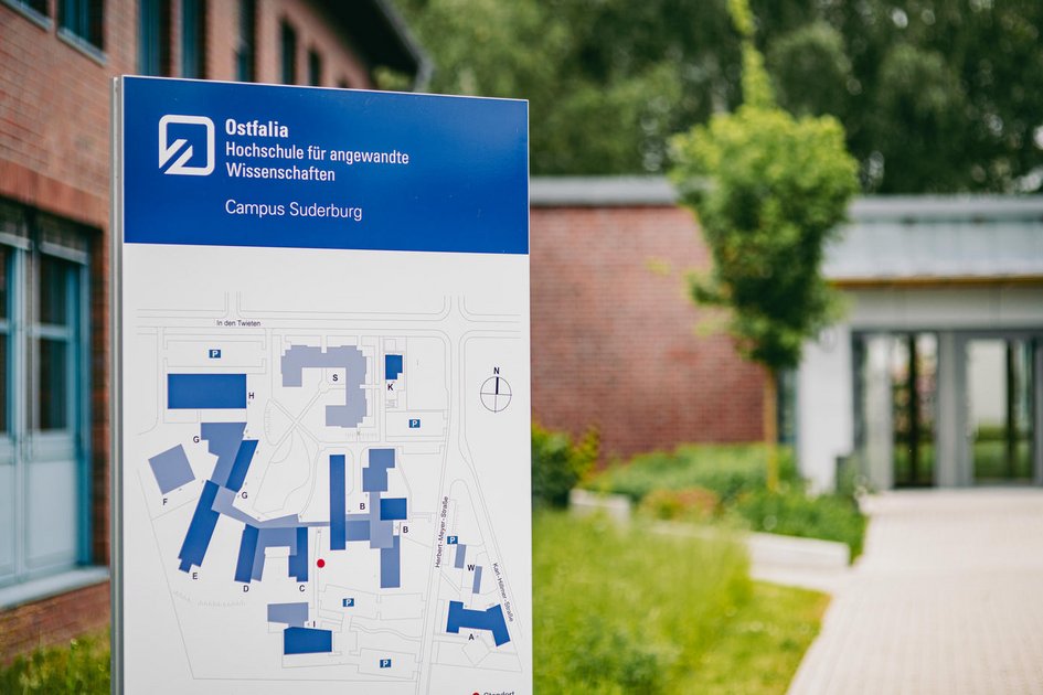 Information sign of the Ostfalia University of Applied Sciences at the Suderburg campus with a campus map in the foreground; a modern building entrance and green areas can be seen in the background.