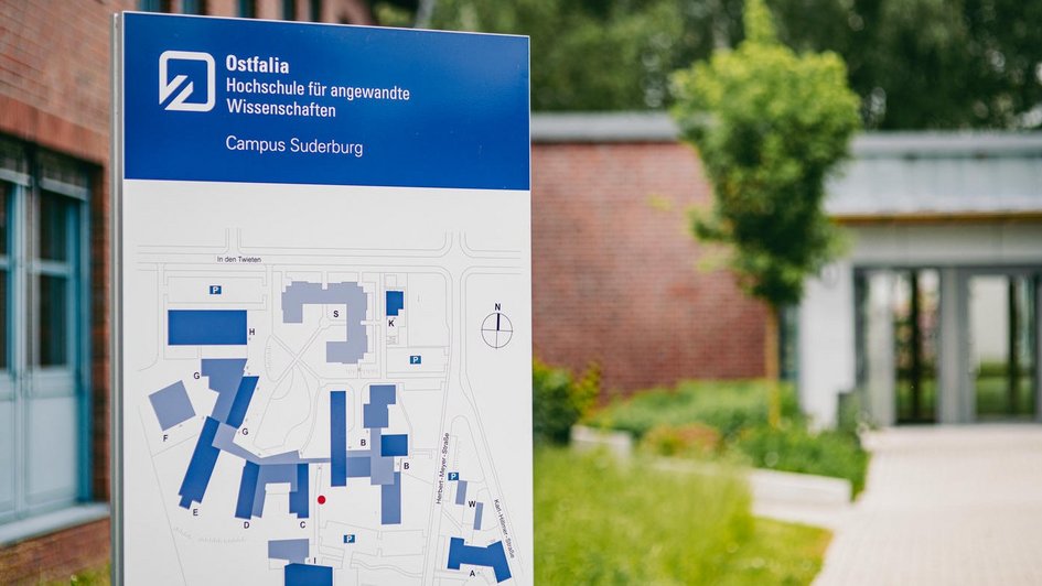 Dekorativ: Informationsschild der Ostfalia Hochschule für angewandte Wissenschaften am Campus Suderburg mit Campusplan im Vordergrund; im Hintergrund ist ein moderner Gebäudeeingang sowie Grünflächen zu sehen. (öffnet Vergrößerung des Bildes)