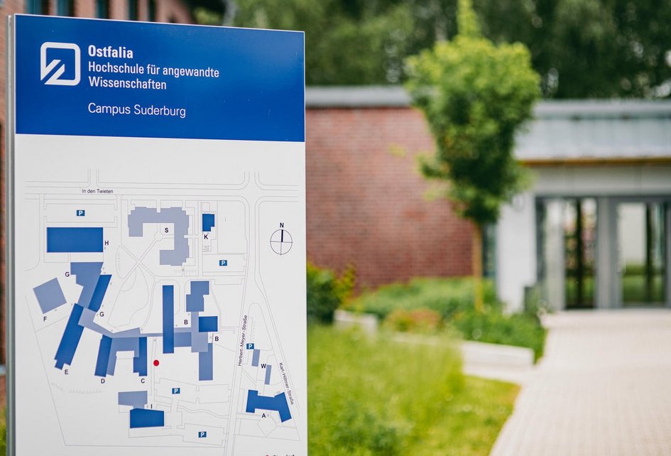 Decorative: Information sign for Ostfalia University of Applied Sciences at the Suderburg campus with a campus map in the foreground; a modern building entrance and green spaces can be seen in the background.