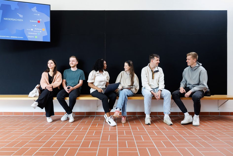 Studierende sitzen auf einer Bank vor einem Hörsaal. Oben links sind auf einem Monitor, die Ostfalia-Standorte auf einer Karte von Niedersachsen zu sehen.