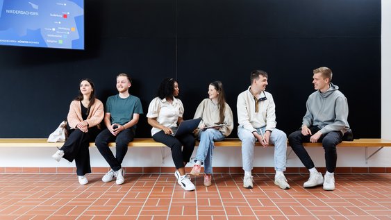 Studierende sitzen auf einer Bank vor einem Hörsaal. Oben links sind auf einem Monitor, die Ostfalia-Standorte auf einer Karte von Niedersachsen zu sehen.