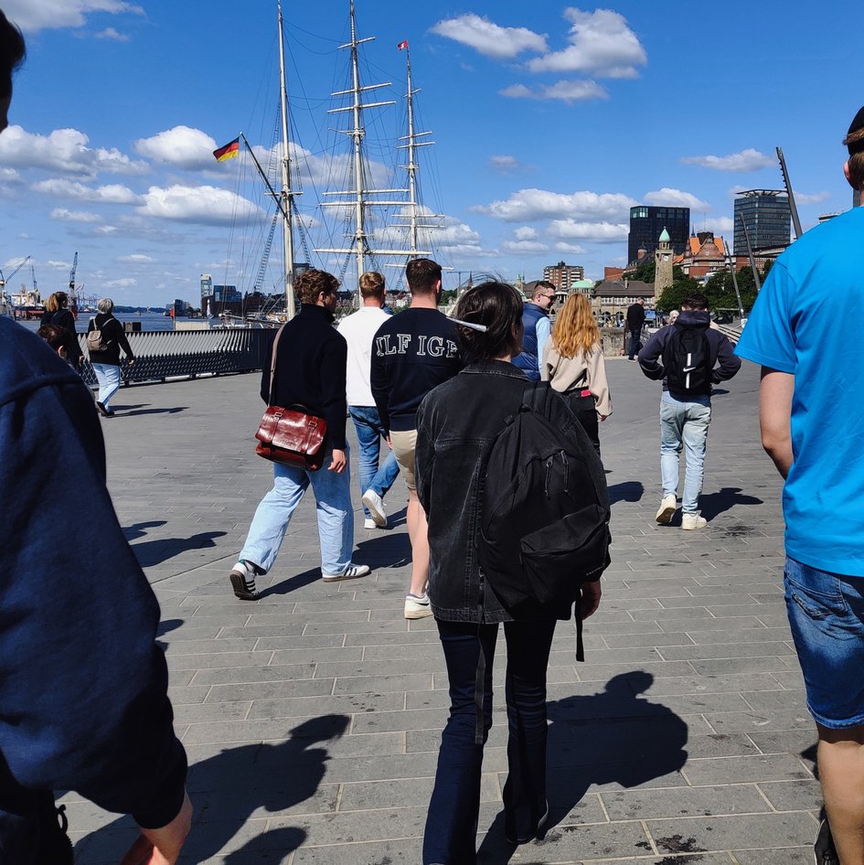 Studierende des 4. Semesters bei einer Exkursion in der Hamburger Hafencity. Im Hintergrund sind die Masten eines Segelschiffes zu sehen.