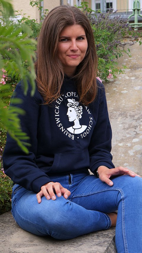 Eine junge Frau trägt den Hoodie der BELS in blau. Der Hoodie trägt das Logo der BELS, die Aequitas, über dem Bauch. Darum findet sich der kreisförmig angeordnete Schriftzug "Brunswick European Law School". Das Logo und der Schriftzug sind weiß. (öffnet Vergrößerung des Bildes)