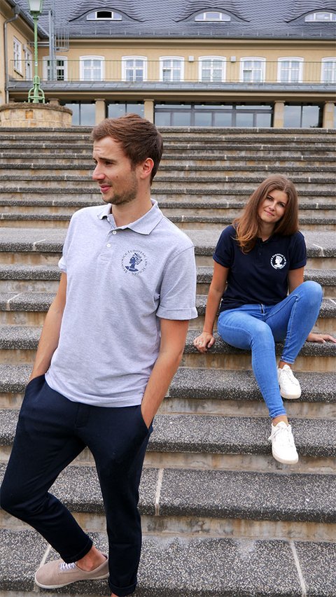 Eine junge Frau trägt das Polo-Shirt der BELS in blau. Ein junger Mann trägt das Polo-Shirt in grau melange. (öffnet Vergrößerung des Bildes)