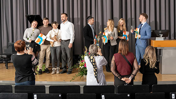 Blick auf die Bühne, vor der Angehörige stehen, welche Fotos von den Absolvent:innen machen, die ihre Urkunden halten.