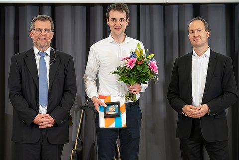 Prof. Dr. Litschen (links), der Preisträger des PKF Fasselt Awards 2025, Carl Thaler, und Sören Damerau von PKF Fasselt. Der Preisträger im weißen Hemd hält seine Auszeichnung und einen bunten Blumenstrauß. Die Abgebildeten lächeln in die Kamera. (öffnet Vergrößerung des Bildes)