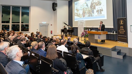 Übersichtsaufnahme mit Blick auf die Bühne, im Vordergrund das Auditorium, am Pult rechts steht Prof. Dr. Litschen, Der Klavierspieler sitzt am Flügel, der links auf der Bühne steht.