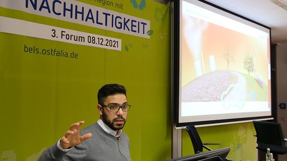 Abbas Seifeddine hält seinen Vortrag beim 3. Forum Nachhaltigkeit. Er steht am Rednerpult, hinter ihm ist eine Leinwand, auf der eine Präsentation gezeigt wird, zu sehen.