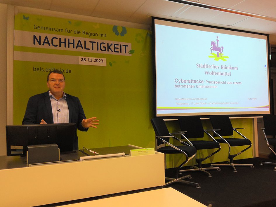 Volker Burghardt erläutert den Ablauf der Cyberattacke auf das Klinikum Wolfenbüttel. Er steht am Rednerpult, auf der Leinwand ist eine Präsentation zu sehen.