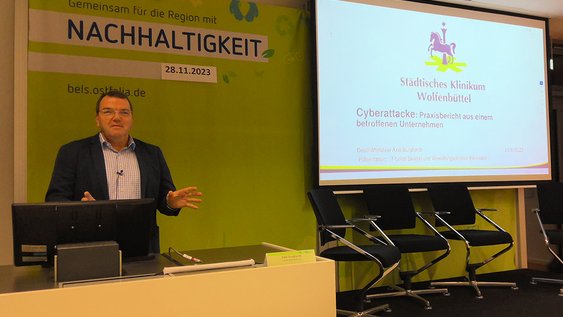 Volker Burghardt erläutert den Ablauf der Cyberattacke auf das Klinikum Wolfenbüttel. Er steht am Rednerpult, auf der Leinwand ist eine Präsentation zu sehen.