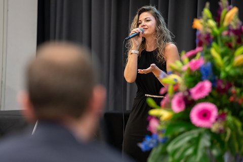 Ina Hallmann, Sängerin des Musikduos Perceived as One trägt ein schwarzes Kleid und singt ins Mikrofon, rechts am Rand des Bildes ist das Blumengesteck zu sehen. (öffnet Vergrößerung des Bildes)