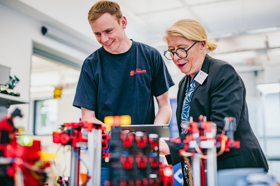 Young man in T-shirt and older woman with glasses and name badge working together on a technical model in the logistics lab.