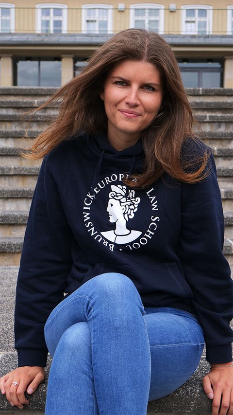 Eine junge Frau trägt den Hoodie der BELS in blau. Der Hoodie trägt das Logo der BELS, die Aequitas, über dem Bauch. Darum findet sich der kreisförmig angeordnete Schriftzug "Brunswick European Law School". Das Logo und der Schriftzug sind weiß. (öffnet Vergrößerung des Bildes)