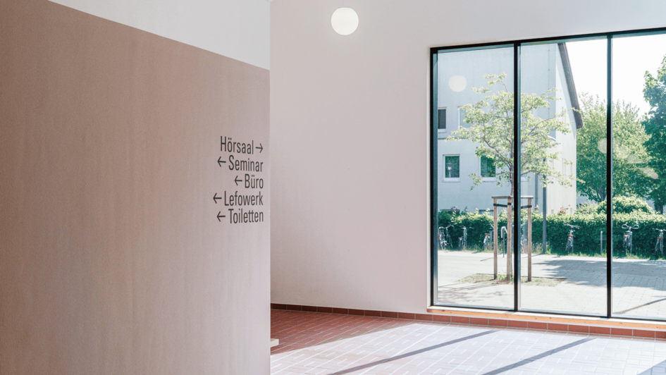Dekorativ:Innenraum mit braunem Fliesenboden, Decke mit Akustikplatten, drei runde Deckenlampen und Wegweiser zu Hörsaal, Seminar, Büro, Labore und Toiletten neben einer großen Glasfront. (öffnet Vergrößerung des Bildes)