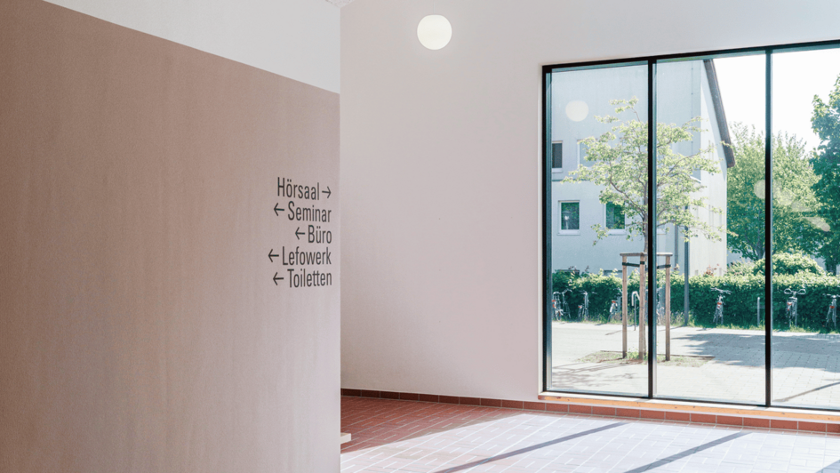 Dekorativ:Innenraum mit braunem Fliesenboden, Decke mit Akustikplatten, drei runde Deckenlampen und Wegweiser zu Hörsaal, Seminar, Büro, Labore und Toiletten neben einer großen Glasfront (öffnet Vergrößerung des Bildes)