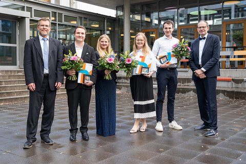 Gruppenbild mit den vier Preisträgerinnen und Preisträgern sowie Dekan und Studiendekan. Die Abgebildeten lächeln in die Kamera. Die Ausgezeichneten halten ihre Awards und Blumensträuße. (öffnet Vergrößerung des Bildes)