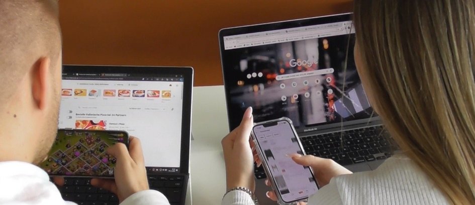 Zu sehen sind ein Student und eine Studentin von hinten. Sie sitzen vor zwei Laptops. Beide halten ein Smartphone in der Hand und tippen.
