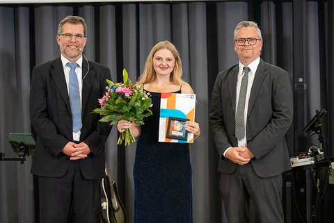 Prof. Dr. Litschen (links), die Preisträgerin des Eckert & Kollegen Awards 2025, Cindy Anton, und Marc Schnieder von Eckert & Kollegen Rechtsanwälte. Die Preisträgerin im dunkelblauen Kleid hält ihre Auszeichnung und einen bunten Blumenstrauß. Die Abgebildeten lächeln in die Kamera. (öffnet Vergrößerung des Bildes)