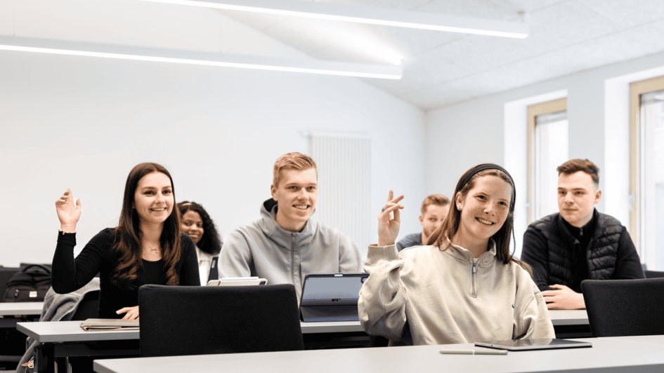 Dekorativ:Kleine Gruppe von Studierenden sitzt in einem hellen Klassenraum, einige heben die Hand, vorne liegen Tablets auf den Tischen (öffnet Vergrößerung des Bildes)