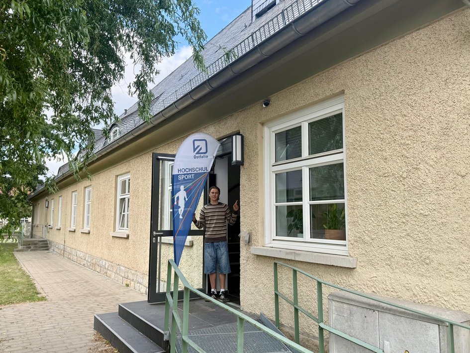 Seiteneingang des Gebäudes Am Exer 3b mit einem Aufsteller „Ostfalia Hochschulsport“. Eine Person steht freundlich winkend in der geöffneten Tür. Der Zugang erfolgt über eine Außentreppe mit Handlauf – nicht barrierefrei.