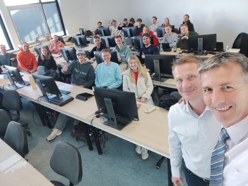 Zwei Kursleiter machen ein Selfie mit ihren Teilnehmenden. Letztere sitzen neben- und hintereinander an Tischen mit Computern