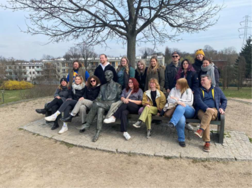 Personen sitzen auf und um eine große Bank herum. Zwischen ihnen sitzt die Statue eines Mannes