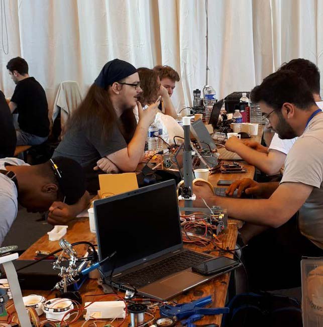 Studenten sitzen am Tisch und arbeiten an Komponenten von Robotern. Zwischen ihnen sind Laptops und Wasserflaschen platiert