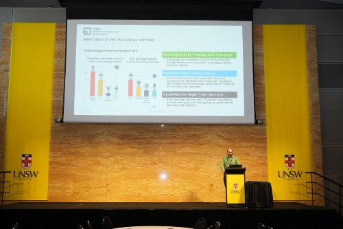 Prof. Stechert steht auf einer Bühne. Eine Präsentation hinter ihm zeigt Grafiken und Balken