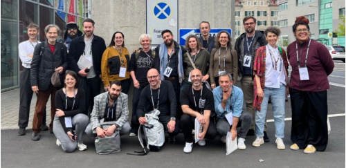 A group of people pose together. The buildings and asphalt of a city can be seen in the background.