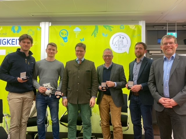 Die Vortragenden vom Forum Nachhaltigkeit stehen vor dem grünen Banner der Veranstaltung und lächeln in die Kamera.