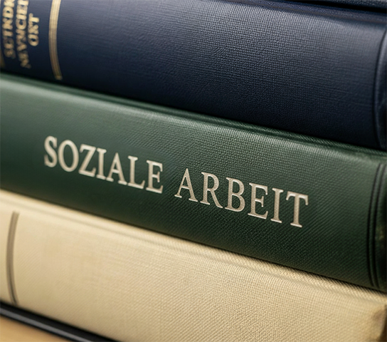 Nahaufnahme eines Stapels gebundener Bücher, bei dem das mittlere, dunkelgrüne Buch im Fokus steht. Auf dessen Buchrücken ist in weißen Buchstaben der Titel "SOZIALE ARBEIT" deutlich zu lesen.